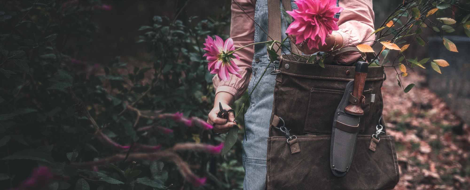 Person wearing a gathering bag with tools and a pink flower, standing in a natural setting.