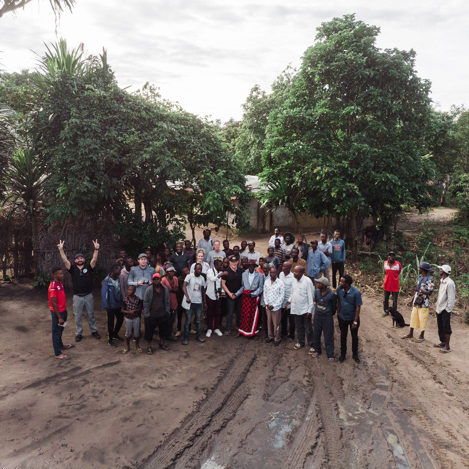 Group of people standing on a dirt road with trees and buildings in the background